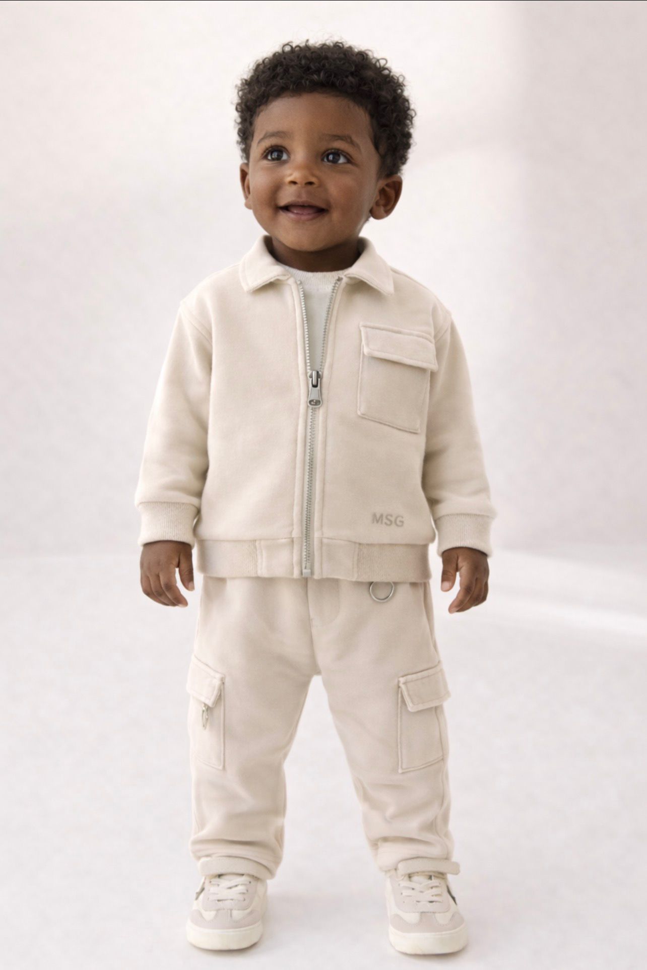 Crèmekleurige jongensset op een jong kind, met een zip jacket en ontspannen broek in een zachte studiosetting.
